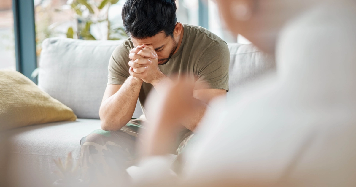 A man sitting on a couch with his hands clasped and head bowed, appearing distressed during a therapy or counseling session, symbolizing PTSD or emotional struggle in Sandy UT